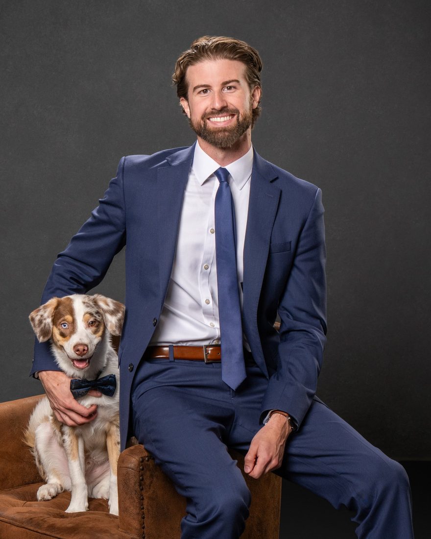 Aidan Suttlehan smiling in a navy suit, seated beside his dog, who is wearing a bow tie